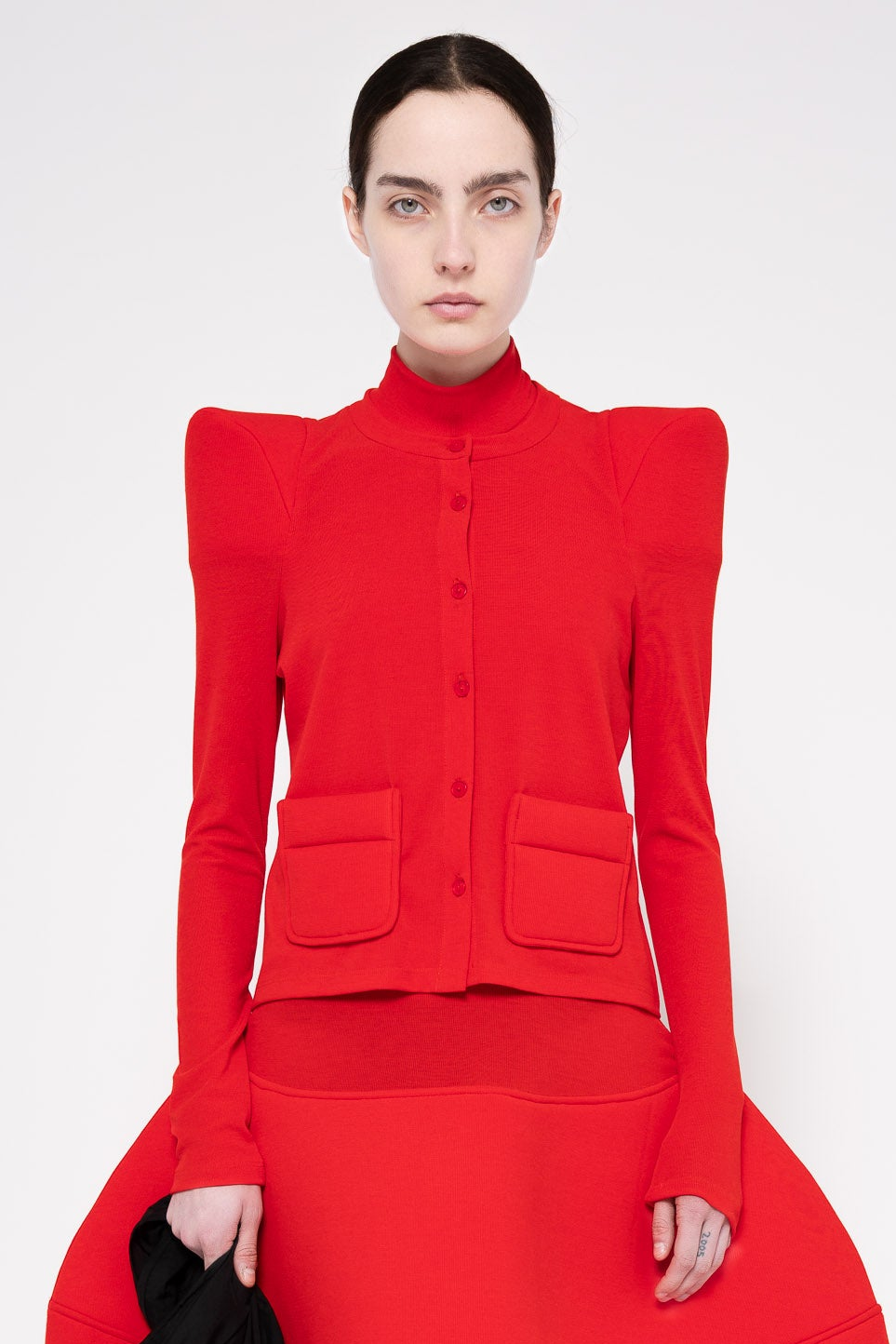 A model wearing a bright red structured knit jacket with dramatic shoulder pads, a high mock neck, front buttons, and two large front patch pockets, paired with a matching red skirt against a light gray studio background.