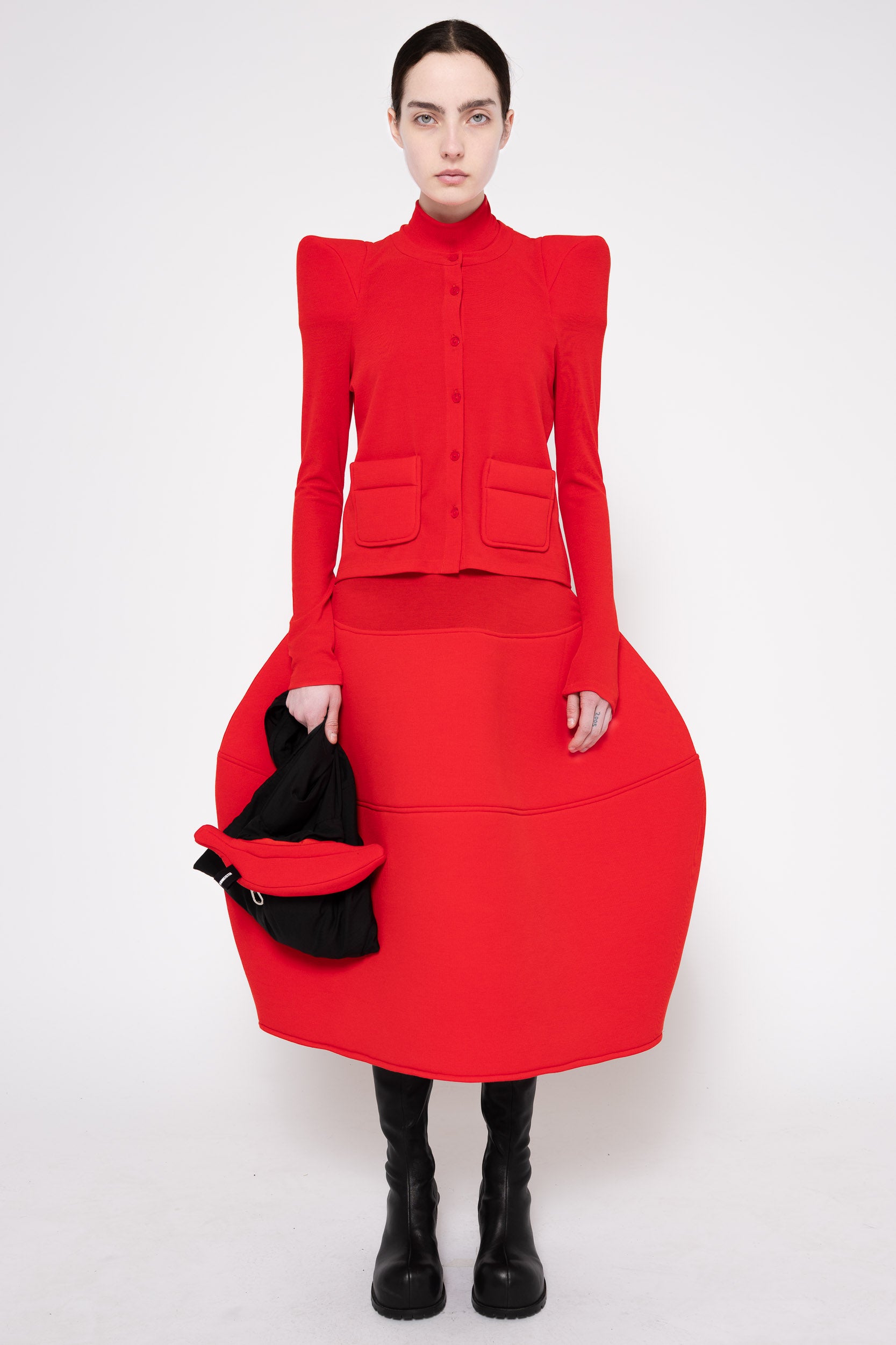 A model stands against a light gray studio background wearing a bright red structured midi dress with dramatic shoulder pads, a high mock neck, front flap pockets, and a very full skirt. She is also wearing black knee-high boots and holding a black hat.