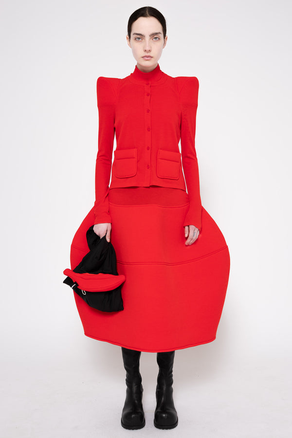 A model stands against a light gray studio background wearing a bright red structured midi dress with dramatic shoulder pads, a high mock neck, front flap pockets, and a very full skirt. She is also wearing black knee-high boots and holding a black hat.
