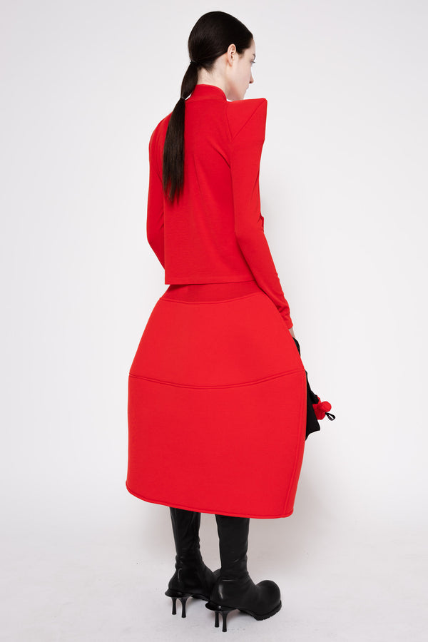 A model shown from the back wearing a bright red two-piece outfit consisting of a cropped long-sleeve top with dramatic angled shoulders and a matching knee-length skirt, paired with black knee-high boots against a plain white studio background.