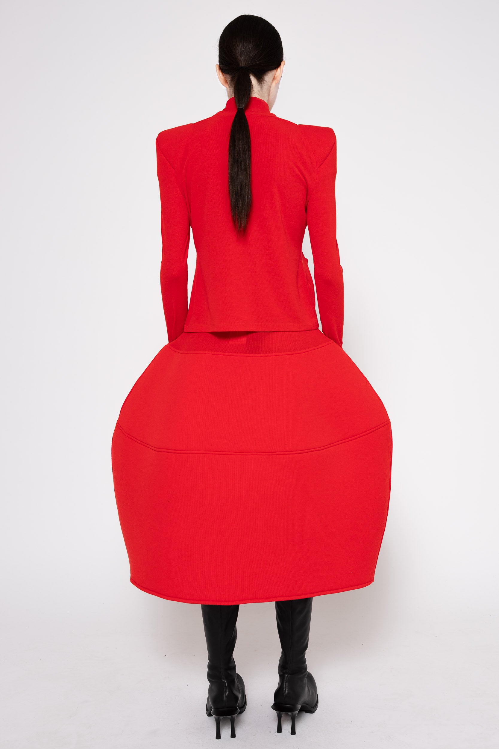 A model shown from the back wearing a bright red structured dress with a high mock neck, padded shoulders, and a voluminous balloon skirt, paired with black boots against a white studio background.