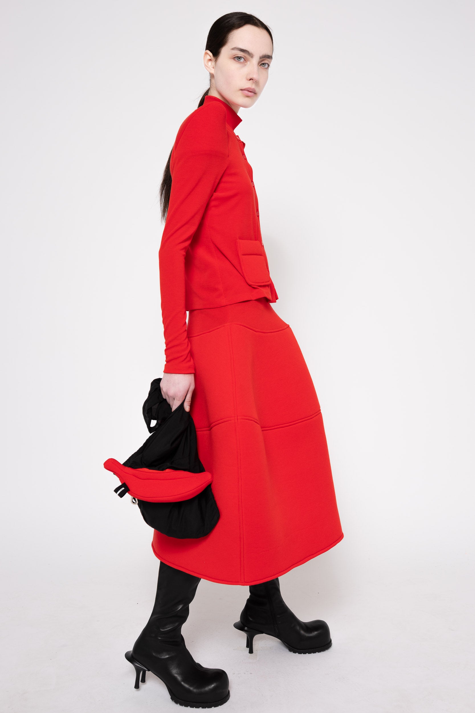 A model wearing a bright red long-sleeve midi dress with a high collar, paired with black heeled boots and holding a black bag, photographed against a plain white studio background.