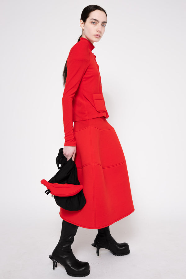 A model wearing a bright red long-sleeve midi dress with a high collar, paired with black heeled boots and holding a black bag, photographed against a plain white studio background.