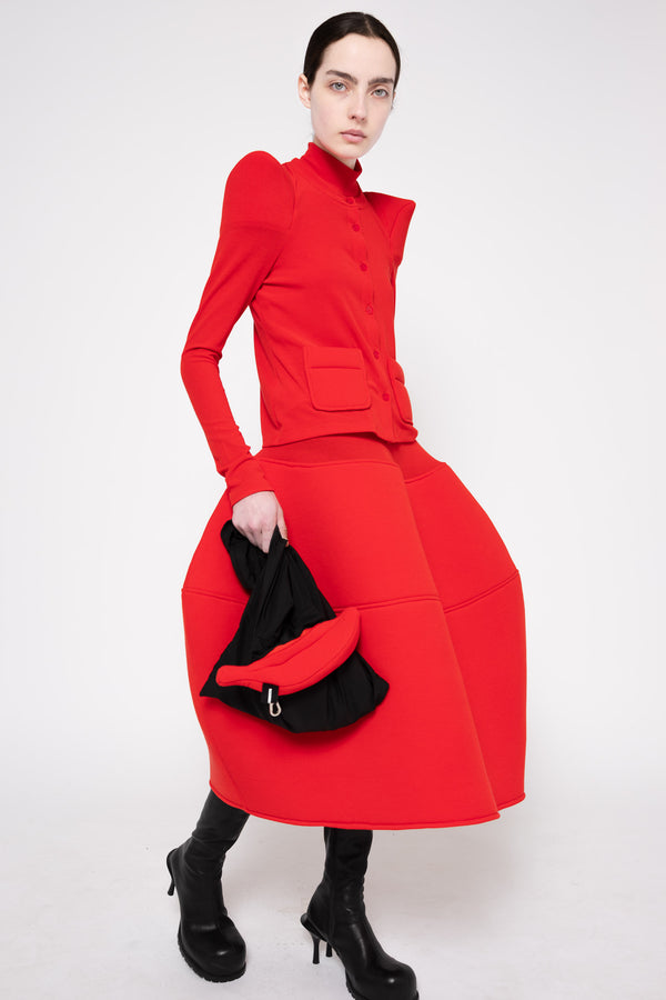 A model wearing a bright red structured midi dress with puffed shoulders, a high mock neck, and a voluminous skirt, holding a black and red handbag. She is wearing black heeled boots against a plain white studio background.