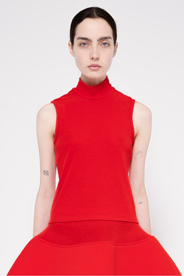 A model wearing a bright red sleeveless mock-neck top and matching red pleated skirt against a light gray studio background.