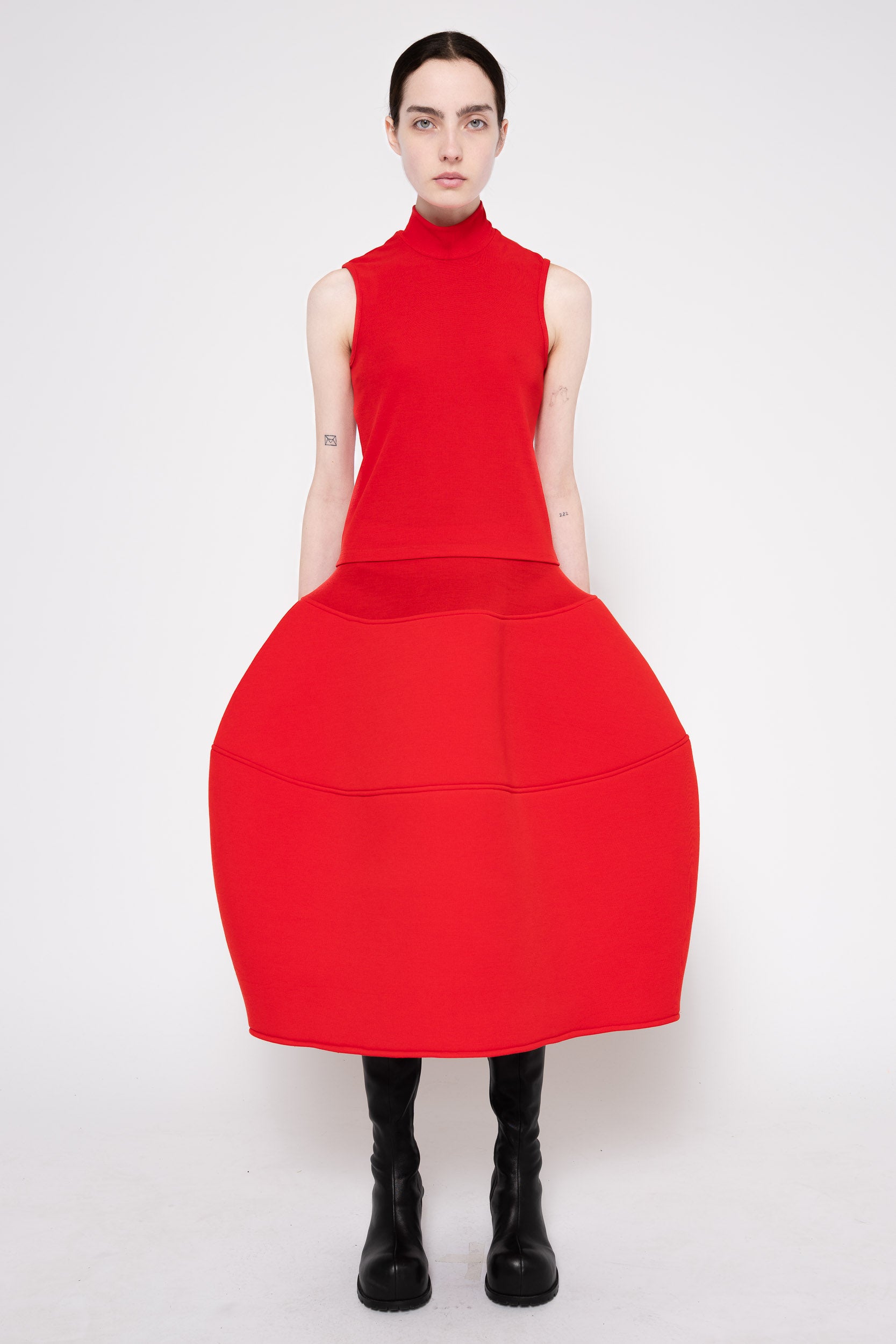 A model wearing a bright red sleeveless mock-neck dress with a very large, structured balloon skirt, paired with black boots, against a plain white studio background.