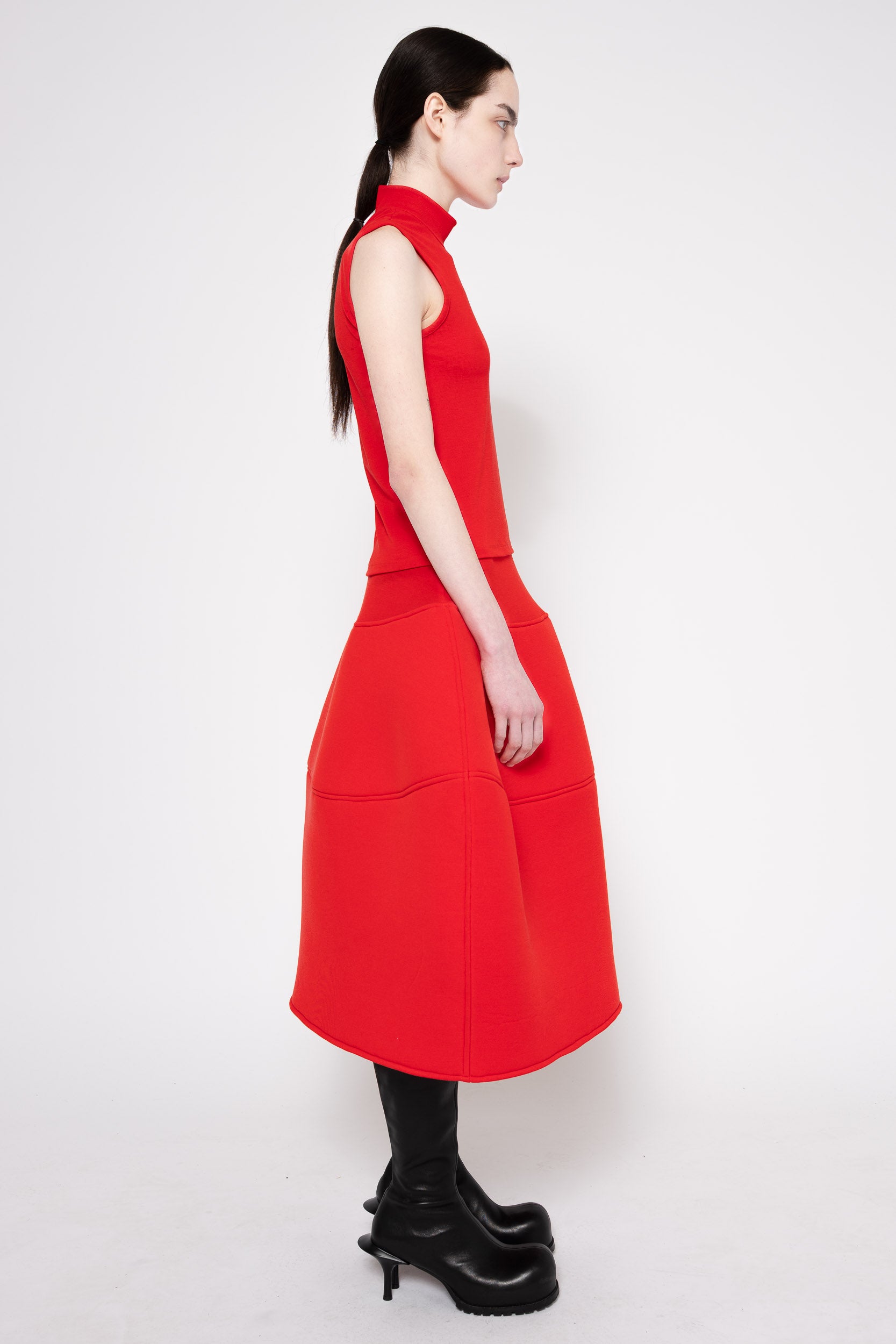 A woman shown from the side wearing a bright red sleeveless midi dress with a structured, balloon-like skirt and black knee-high boots, photographed against a light gray studio background.