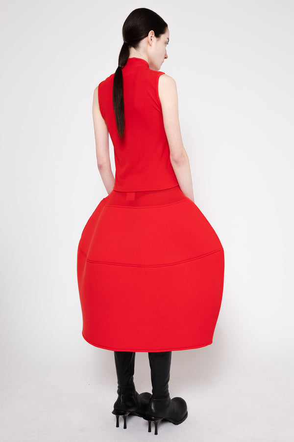A woman shown from the back wearing a bright red sleeveless midi dress with a structured, balloon-like skirt and black knee-high boots, photographed against a light gray studio background.
