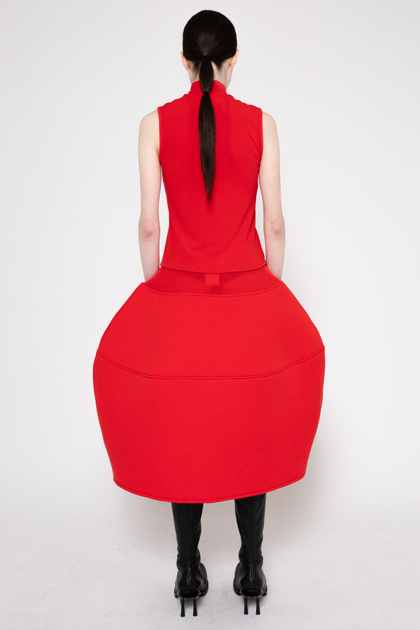 A model shown from the back wearing a bright red sleeveless midi dress with a high mock neck and a very full, balloon-like skirt, paired with black knee-high socks and black high-heeled boots, against a plain white studio background.