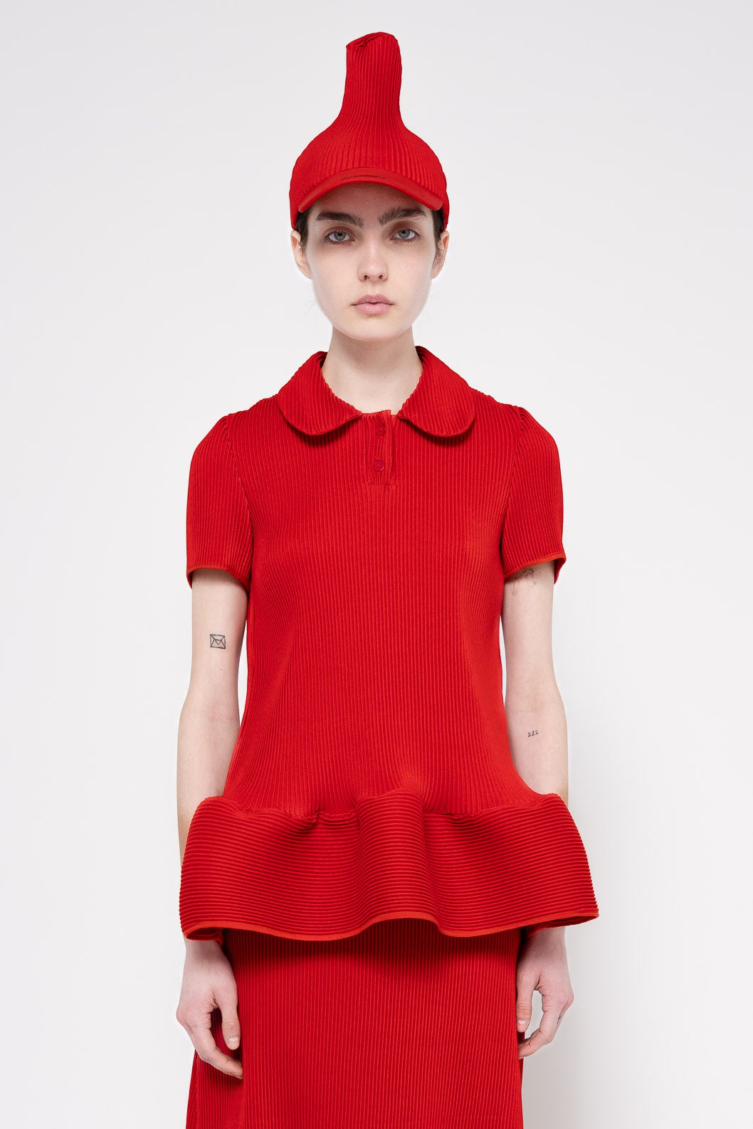 A model wearing a bright red ribbed knit outfit consisting of a short-sleeve collared top with a ruffled hem, matching red skirt, and a pointed red hat, photographed against a light gray studio background.