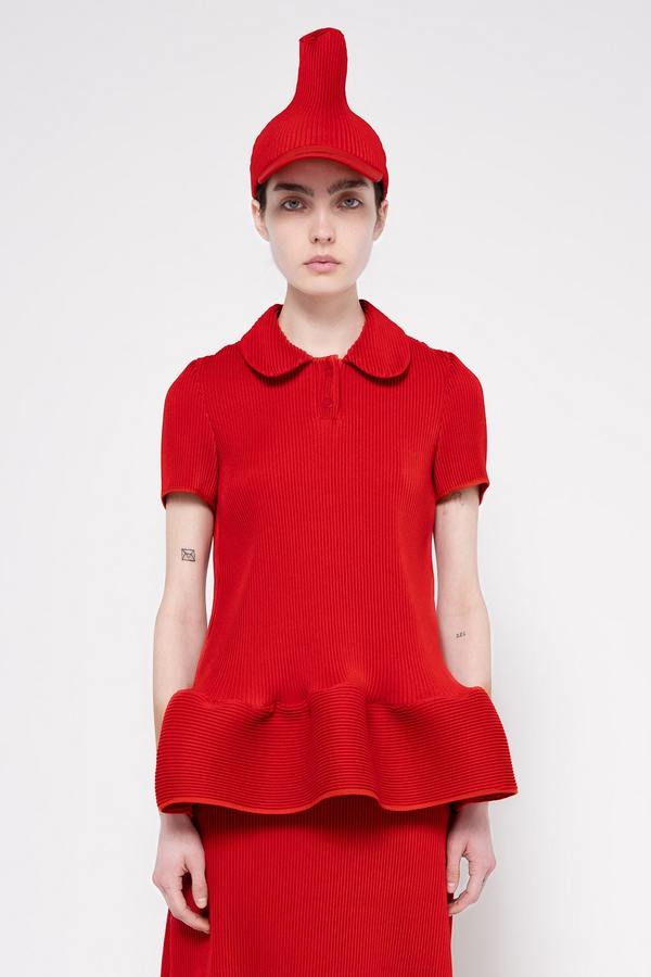 A model wearing a bright red ribbed knit outfit consisting of a short-sleeve collared top with a ruffled hem, matching red skirt, and a pointed red hat, photographed against a light gray studio background.