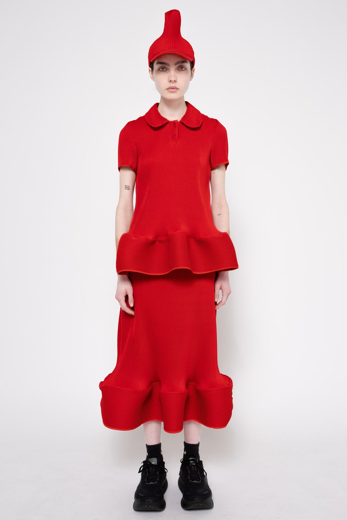 A model stands against a light gray studio background wearing a bright red matching outfit consisting of a short-sleeve collared top with a ruffled waist, a long ruffled skirt, and a pointed red hat. The model is also wearing black socks and black boots.