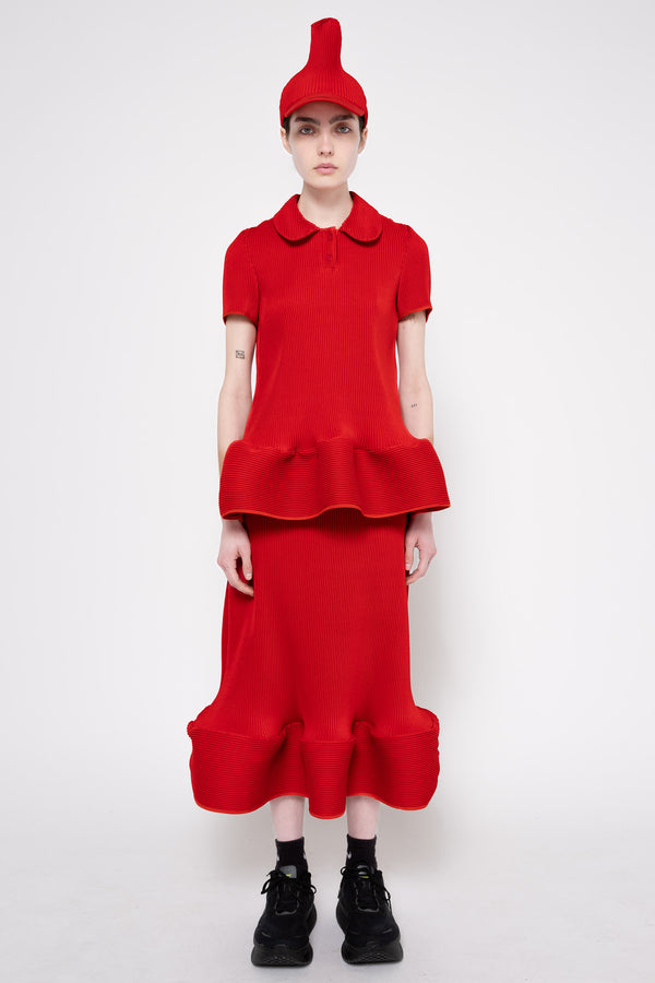 A model stands against a light gray studio background wearing a bright red matching outfit consisting of a short-sleeve collared top with a ruffled waist, a long ruffled skirt, and a pointed red hat. The model is also wearing black socks and black boots.