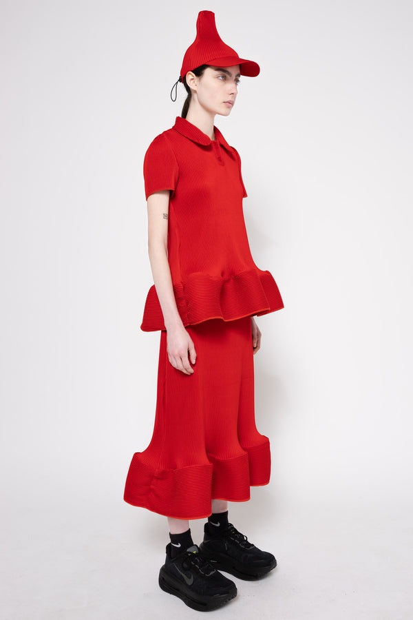 A model wearing a bright red two-piece outfit consisting of a short-sleeve collared top with a ruffled hem and matching wide-leg ruffled pants, paired with a red pointed cap and black sneakers, against a plain light gray studio background.