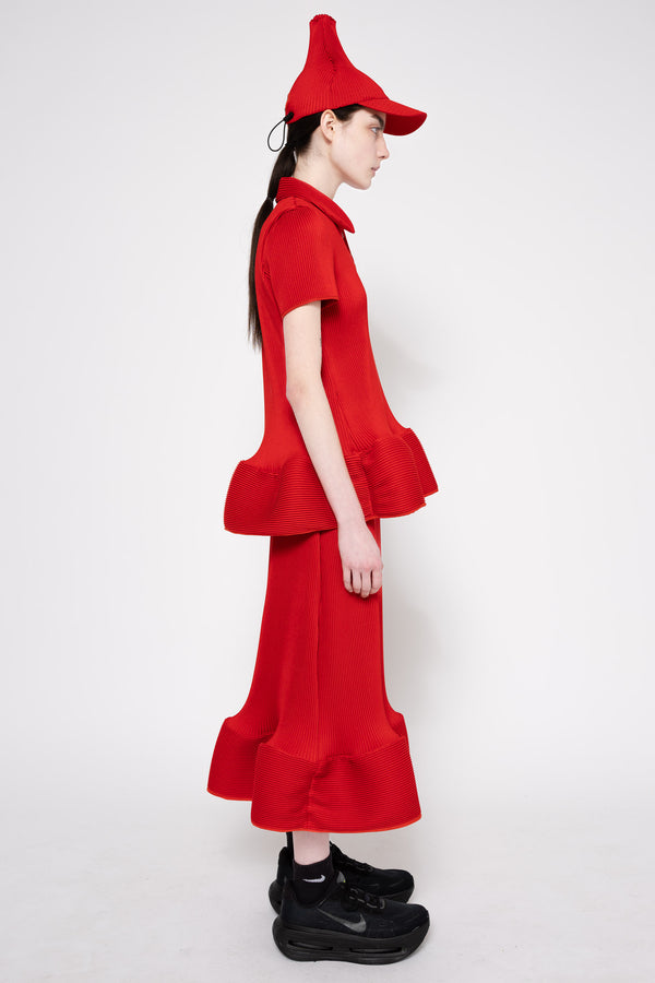 A model in profile wearing a bright red two-piece outfit with a short-sleeve collared top, matching wide-leg skirt, and a pointed red hat, paired with black sneakers, against a plain white studio background.