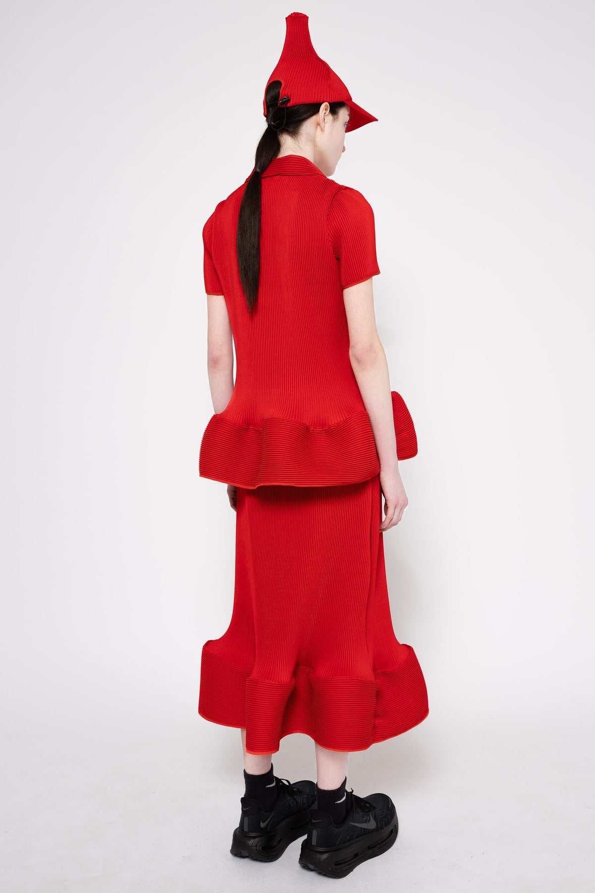A model shown from the back wearing a bright red two-piece outfit consisting of a short-sleeve top with a ruffled hem and a matching long skirt, along with a pointed red hat and black shoes, against a plain light gray background.