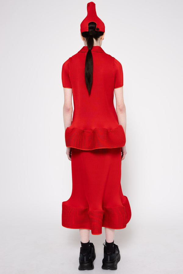 A model shown from the back wearing a bright red two-piece outfit consisting of a short-sleeve top with a ruffled hem and a matching high-waisted flared skirt, paired with a red pointed hat and black boots, against a light gray background.