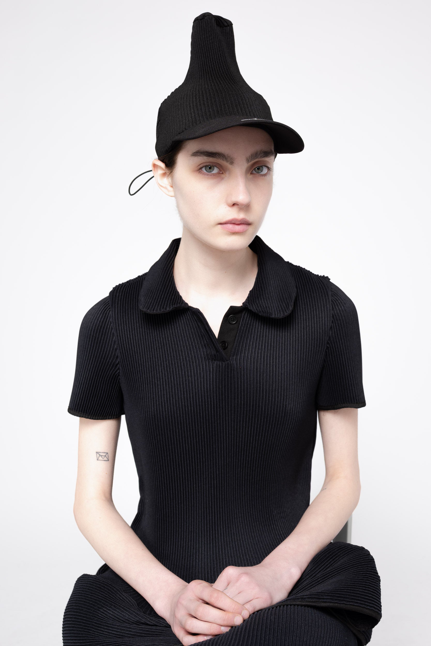 A model wearing a black pleated short-sleeve polo shirt, matching black pleated skirt, and a black pleated hat, posed against a light gray studio background.