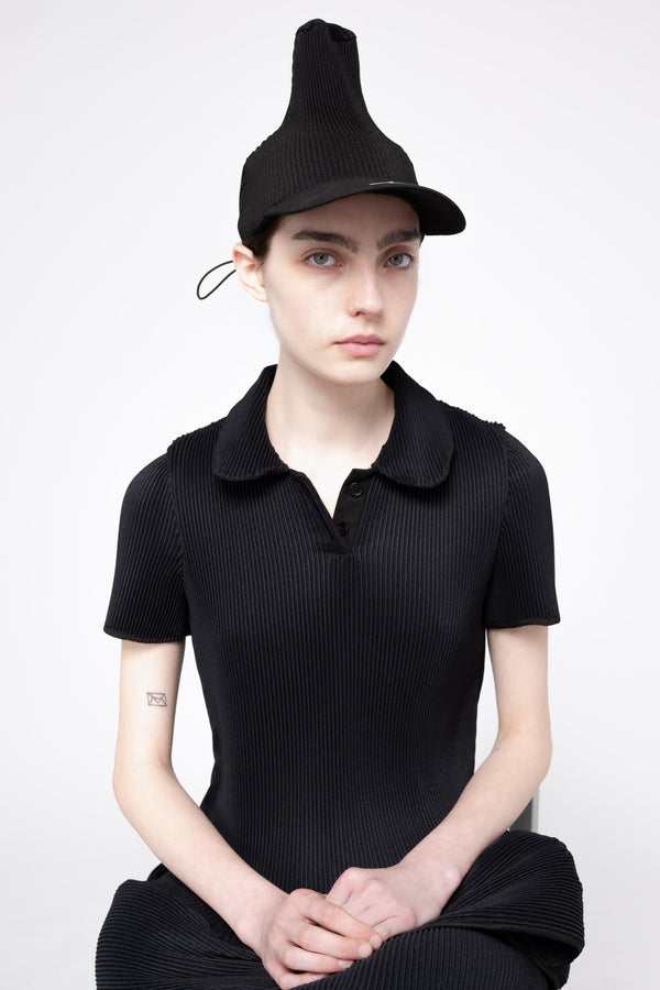 A model wearing a black pleated short-sleeve polo shirt, matching black pleated skirt, and a black pleated hat, posed against a light gray studio background.