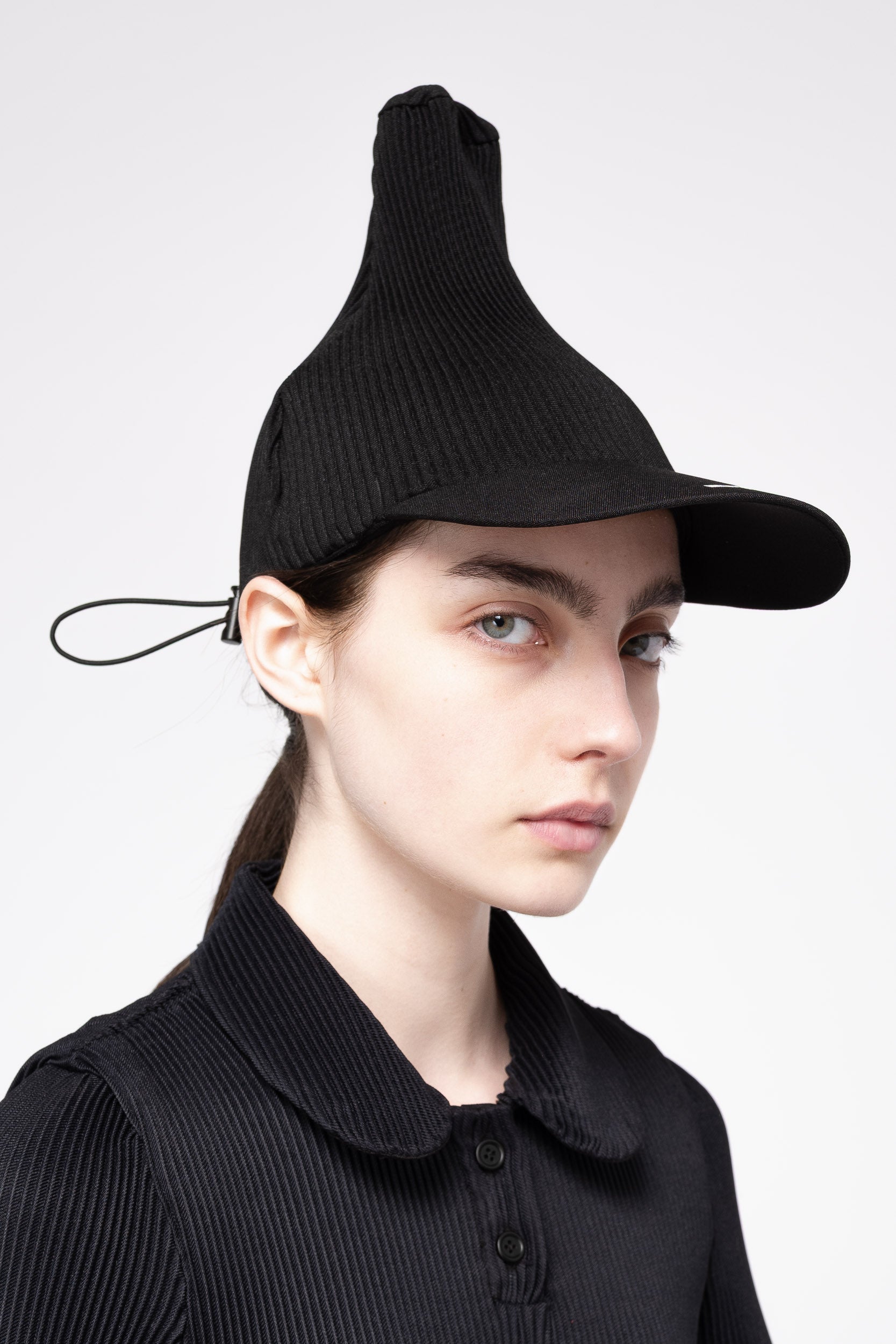 A model wearing a black pleated cap with a unique pointed top and short brim, paired with a matching black ribbed knit polo shirt, against a light gray background.