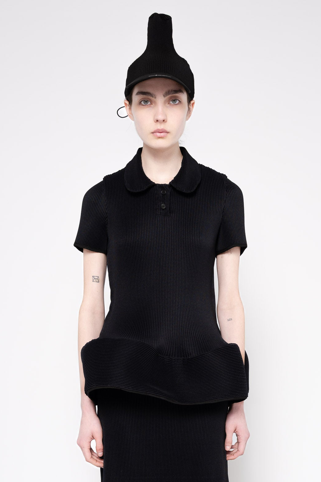 A model wearing a black ribbed short-sleeve polo shirt with a unique folded hem, paired with a matching black beanie and skirt, against a plain light gray background.