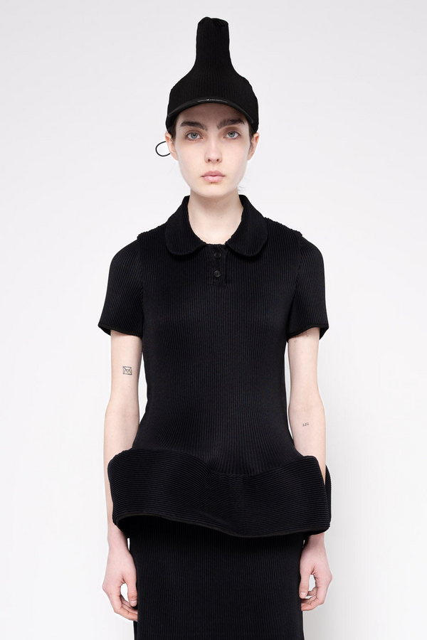 A model wearing a black ribbed short-sleeve polo shirt with a unique folded hem, paired with a matching black beanie and skirt, against a plain light gray background.