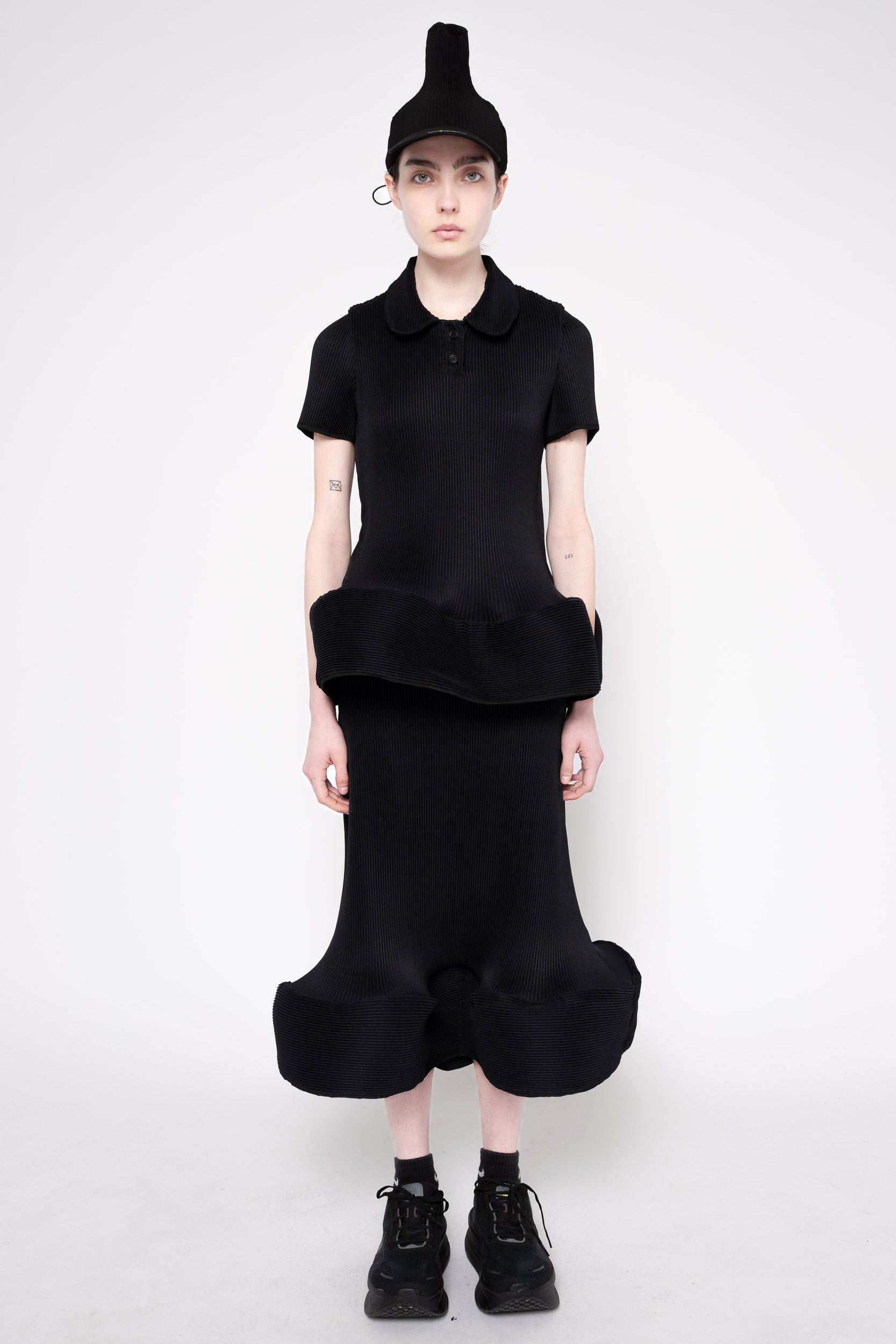 A model wearing a black short-sleeve collared top with a ruffled hem and a matching black voluminous skirt, styled with a black pointed cap and black lace-up shoes against a light gray studio background.