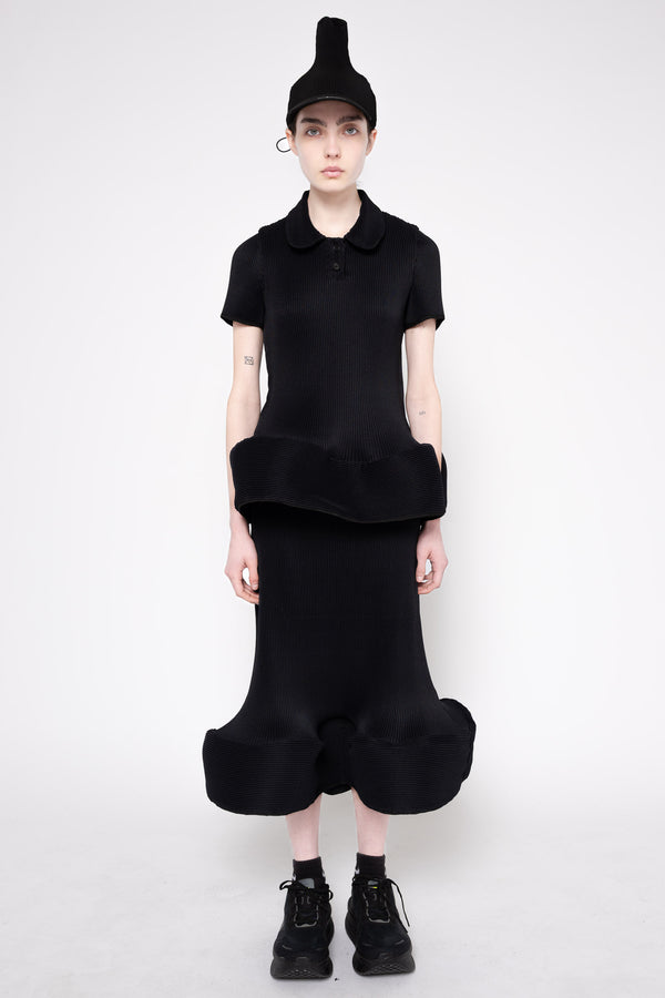 A model wearing a black short-sleeve collared top with a ruffled hem and a matching black voluminous skirt, styled with a black pointed cap and black lace-up shoes against a light gray studio background.