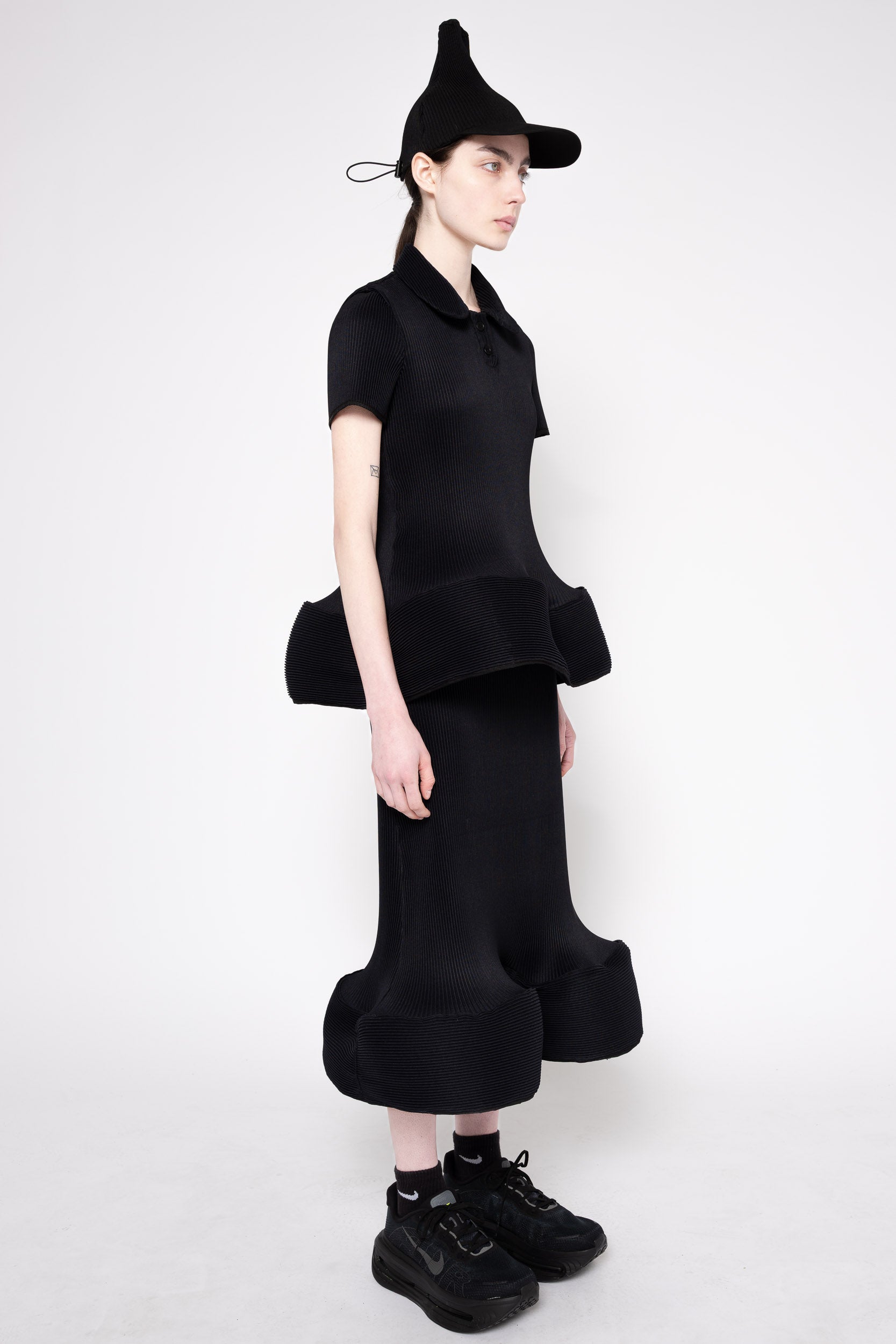 A model wearing a black fashion outfit consisting of a short-sleeve collared top with dramatic ruffled layers, matching wide-leg ruffled pants, black socks, black sneakers, and a pointed black hat, photographed against a light gray studio background.