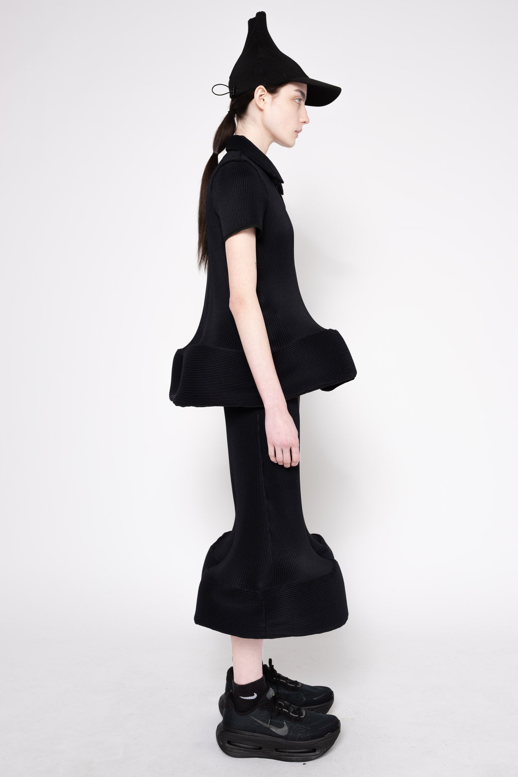 A model in profile wearing a black short-sleeve collared top with a dramatic ruffled hem, matching flared black pants, a pointed black hat, and black sneakers against a plain light gray studio background.
