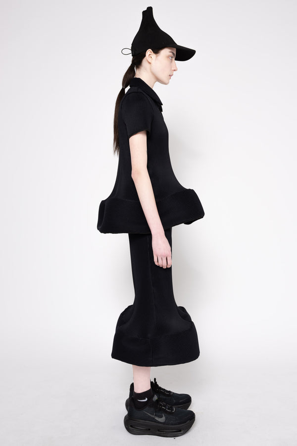 A model in profile wearing a black short-sleeve collared top with a dramatic ruffled hem, matching flared black pants, a pointed black hat, and black sneakers against a plain light gray studio background.