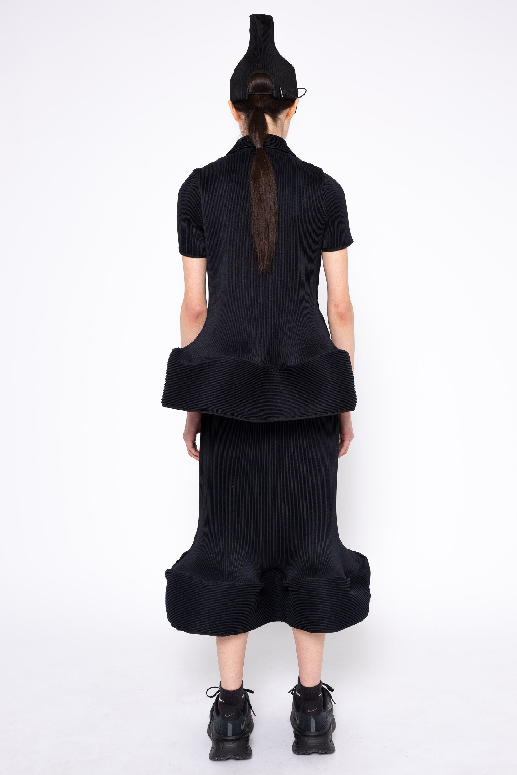 A model viewed from the back wearing a black short-sleeve top with a dramatic ruffled waist and a matching black skirt, paired with black shoes and a black hat, against a plain white studio background.