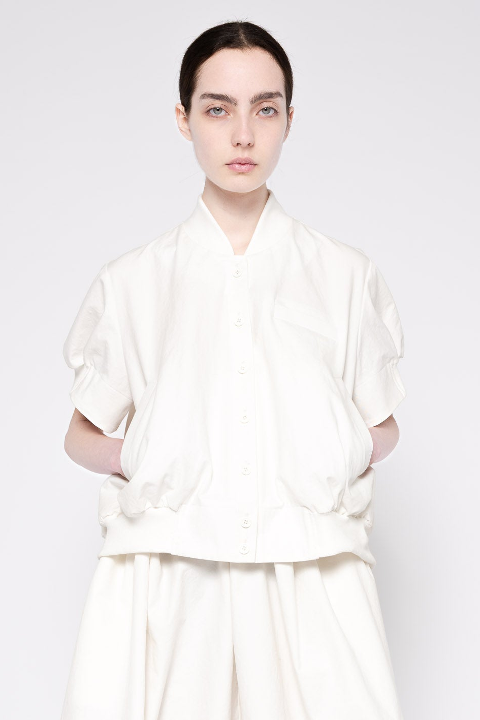 A model wearing a white short-sleeve button-front top with gathered, voluminous sleeves and a relaxed fit, paired with matching white wide-leg pants, against a light gray studio background.