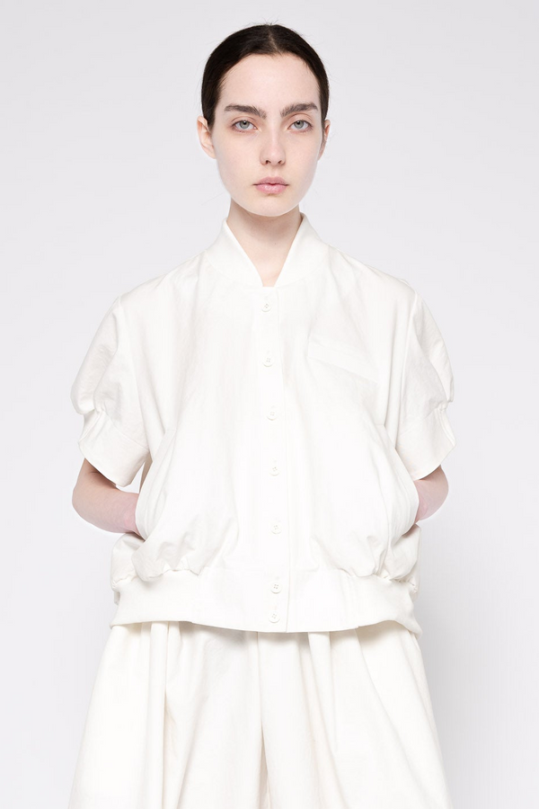 A model wearing a white short-sleeve button-front top with gathered, voluminous sleeves and a relaxed fit, paired with matching white wide-leg pants, against a light gray studio background.
