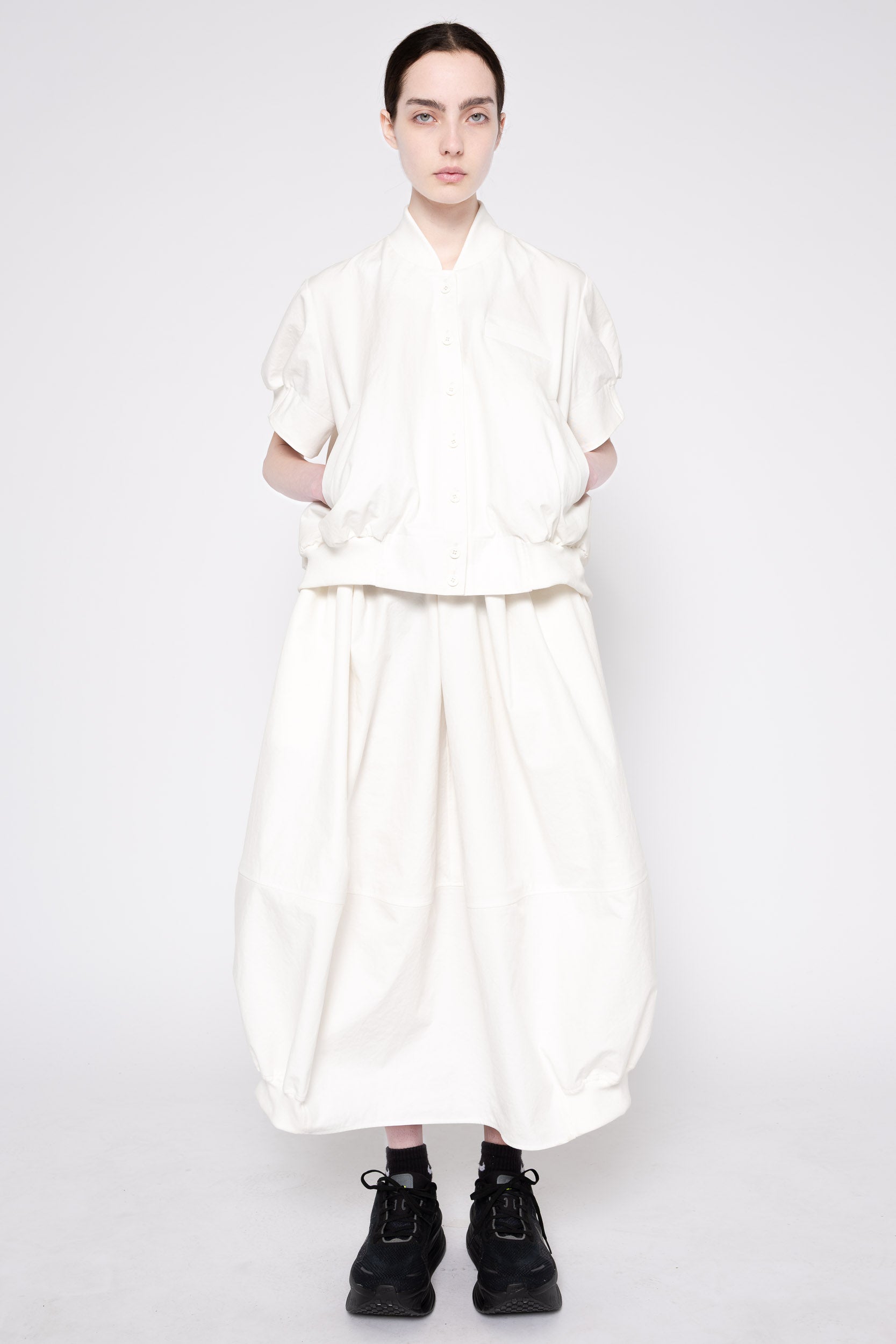 A model wearing a voluminous, off-white two-piece outfit consisting of a short-sleeved top and matching wide-leg pants, paired with black socks and black sneakers, against a plain light gray background.
