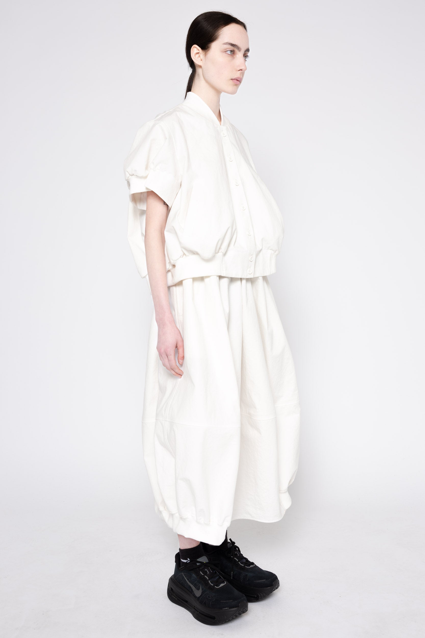 A model wearing a voluminous, off-white two-piece outfit consisting of a short-sleeved top and matching wide-leg pants, paired with black sneakers, against a plain light gray studio background.