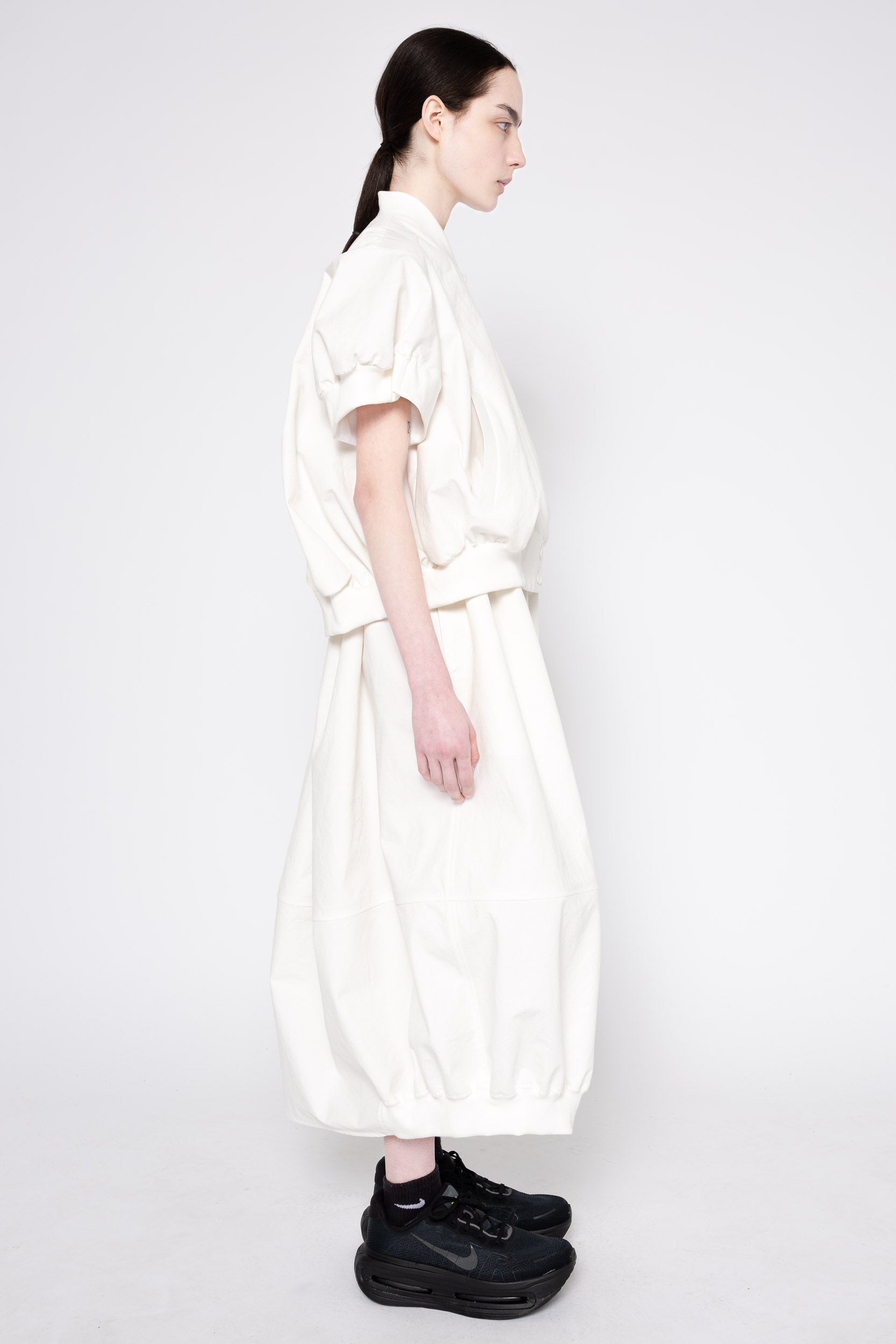 A model shown in profile wearing a loose-fitting, high-collar white dress with voluminous short sleeves and a belted waist, paired with black sneakers, against a plain light gray studio background.