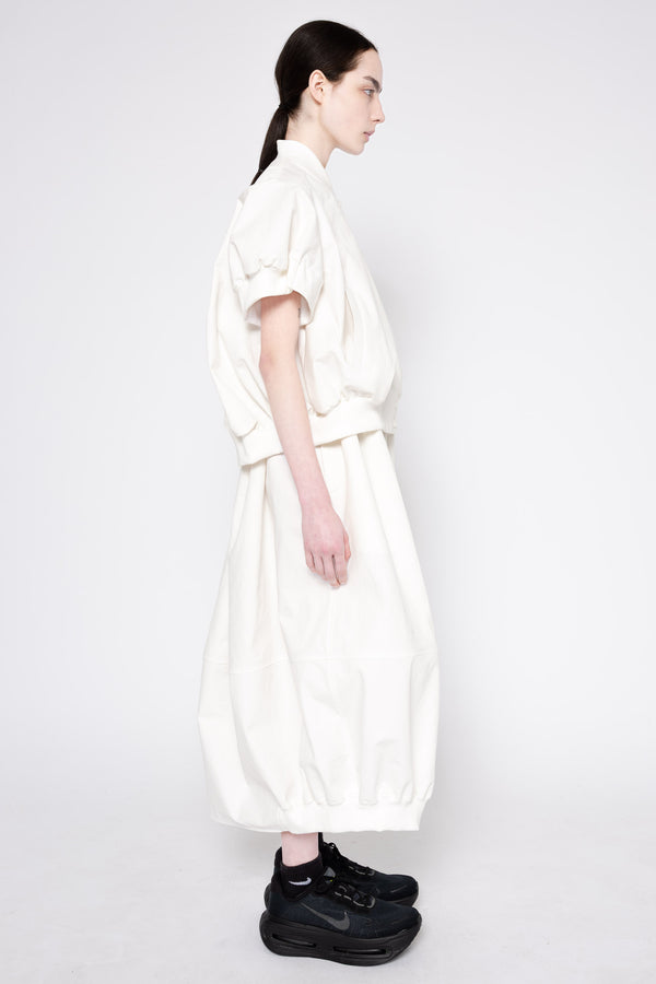 A model shown in profile wearing a loose-fitting, high-collar white dress with voluminous short sleeves and a belted waist, paired with black sneakers, against a plain light gray studio background.