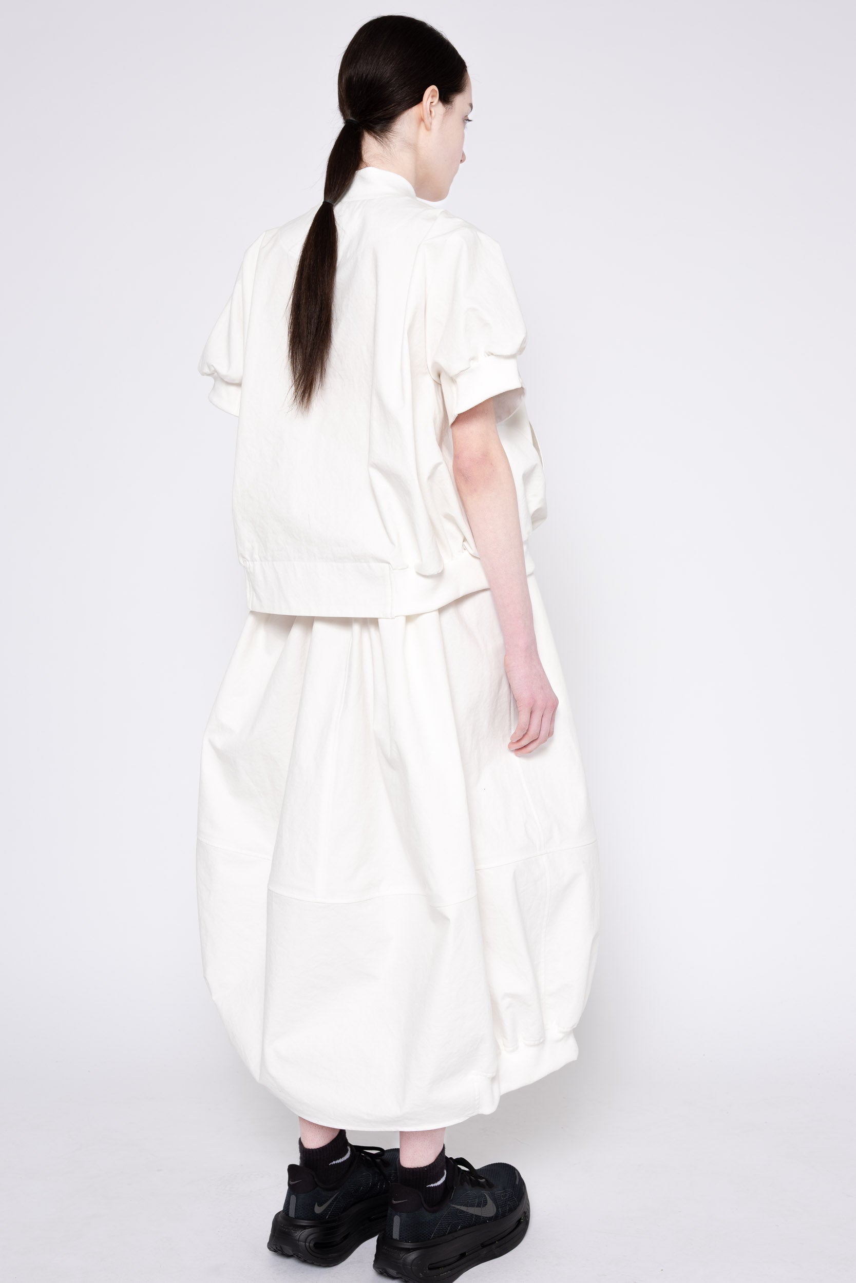 A model shown from the back wearing a white high-neck, short-sleeved cropped jacket and matching wide-leg skirt, paired with black platform sneakers, against a plain white studio background.