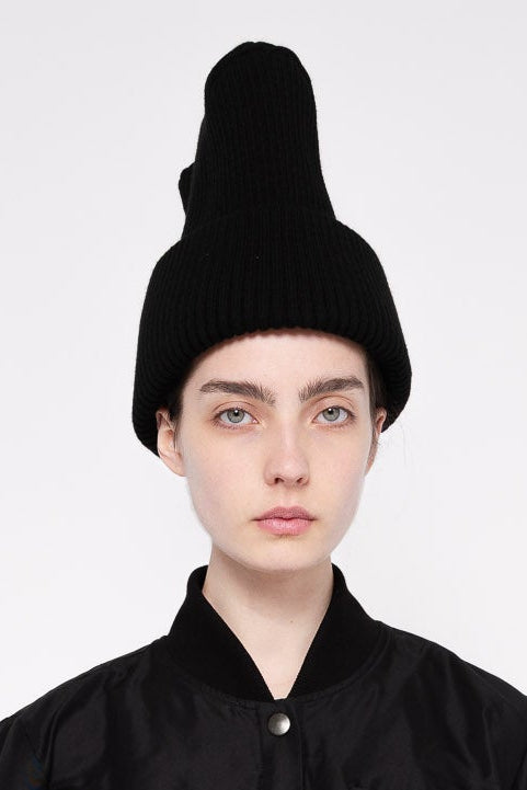 A model wearing a tall, black ribbed knit beanie hat and a black jacket, photographed against a plain light gray background.