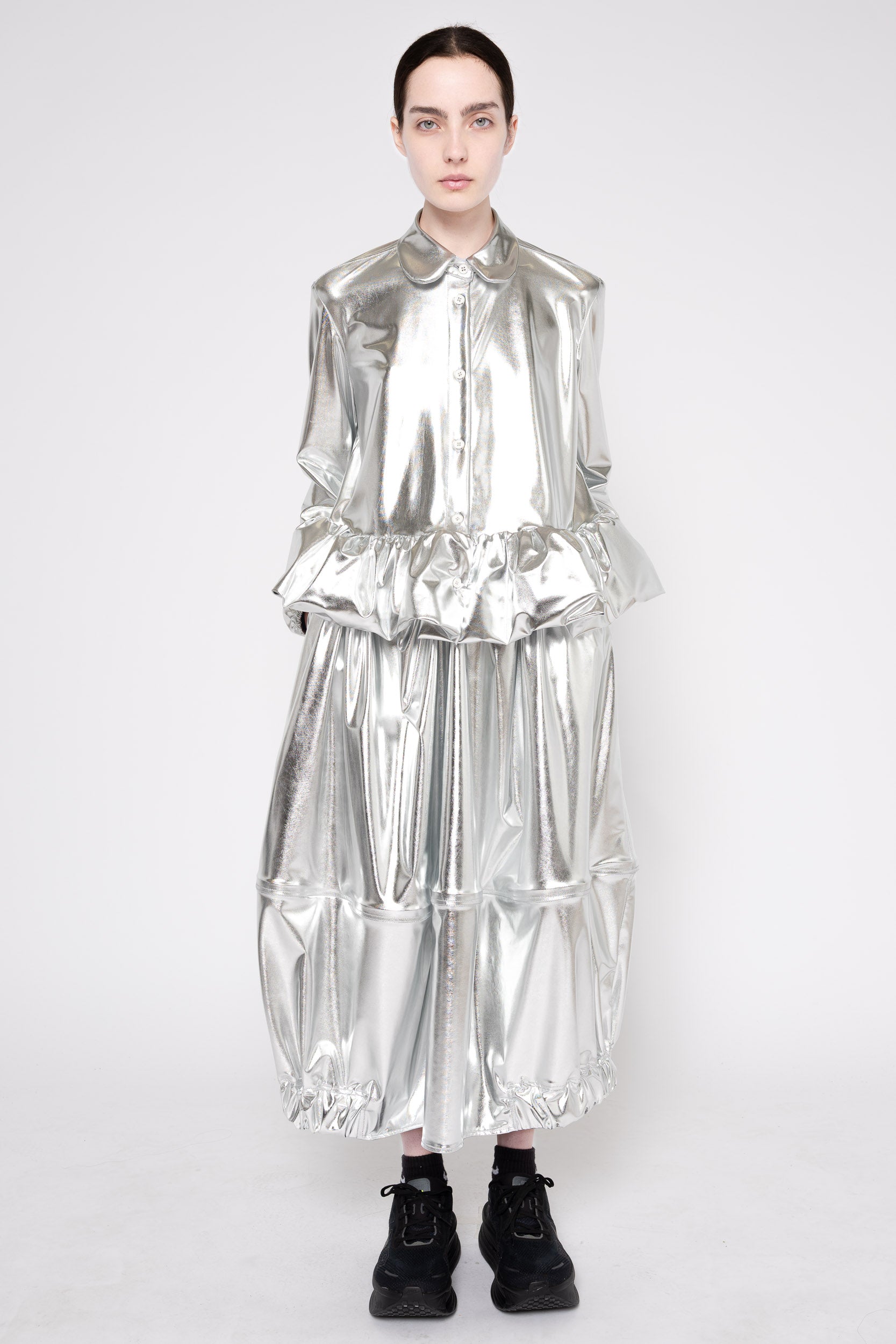 A model stands against a light gray studio background wearing a highly reflective, metallic silver long-sleeve dress with a ruffled collar and voluminous tiered skirt, paired with black lace-up boots.