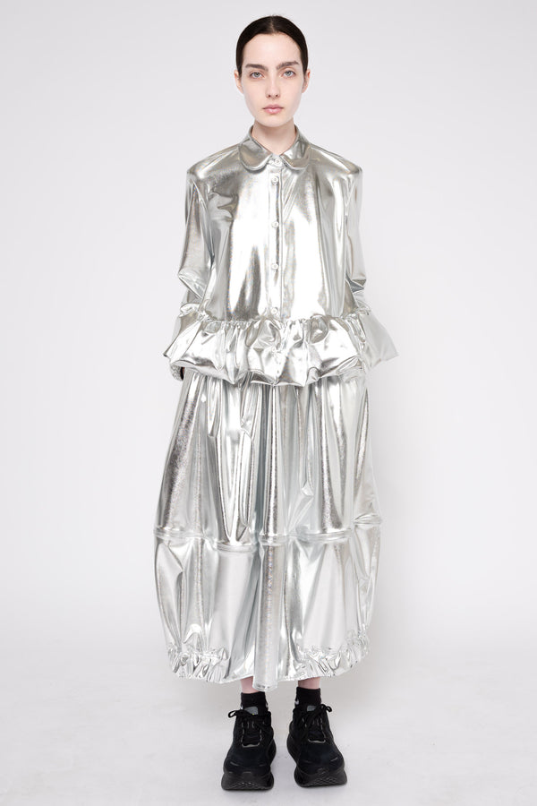 A model stands against a light gray studio background wearing a highly reflective, metallic silver long-sleeve dress with a ruffled collar and voluminous tiered skirt, paired with black lace-up boots.