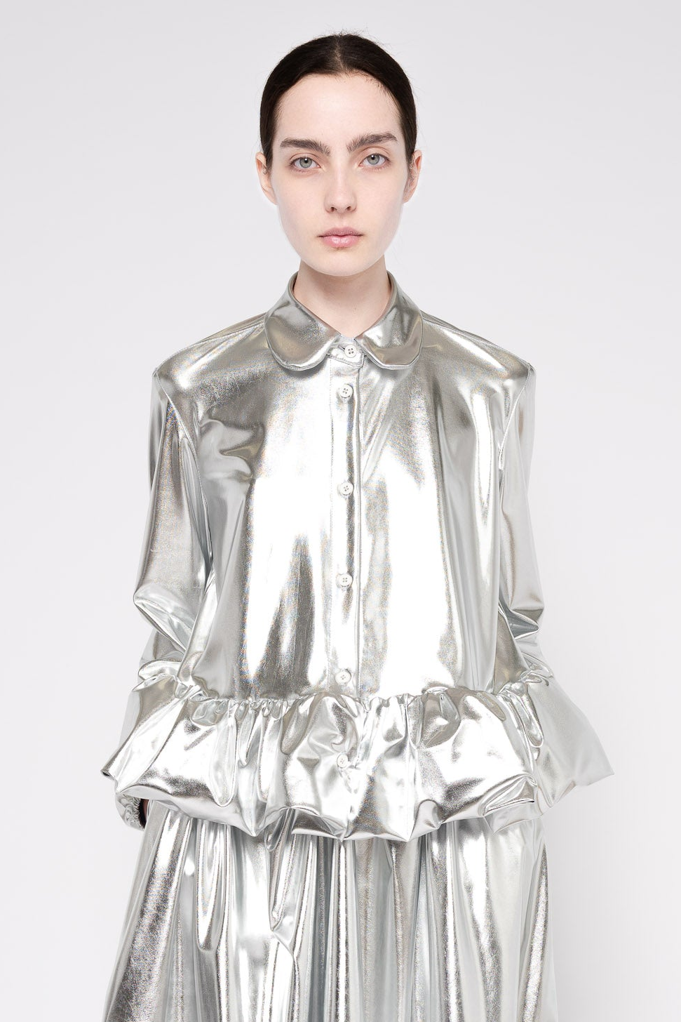 A model wearing a shiny metallic silver long-sleeve blouse with a Peter Pan collar and a matching pleated skirt, photographed against a light gray studio background.