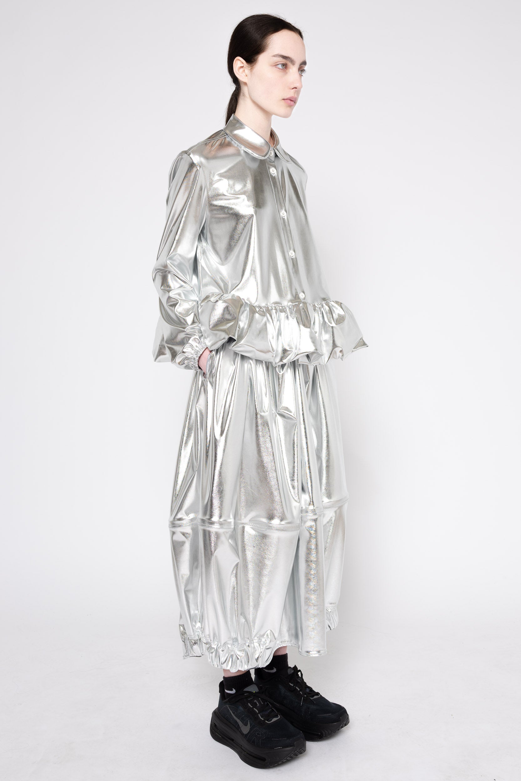 A model wearing a shiny metallic silver long-sleeve dress with a high collar, gathered waist, and voluminous tiered skirt, paired with black sneakers, against a plain light gray background.