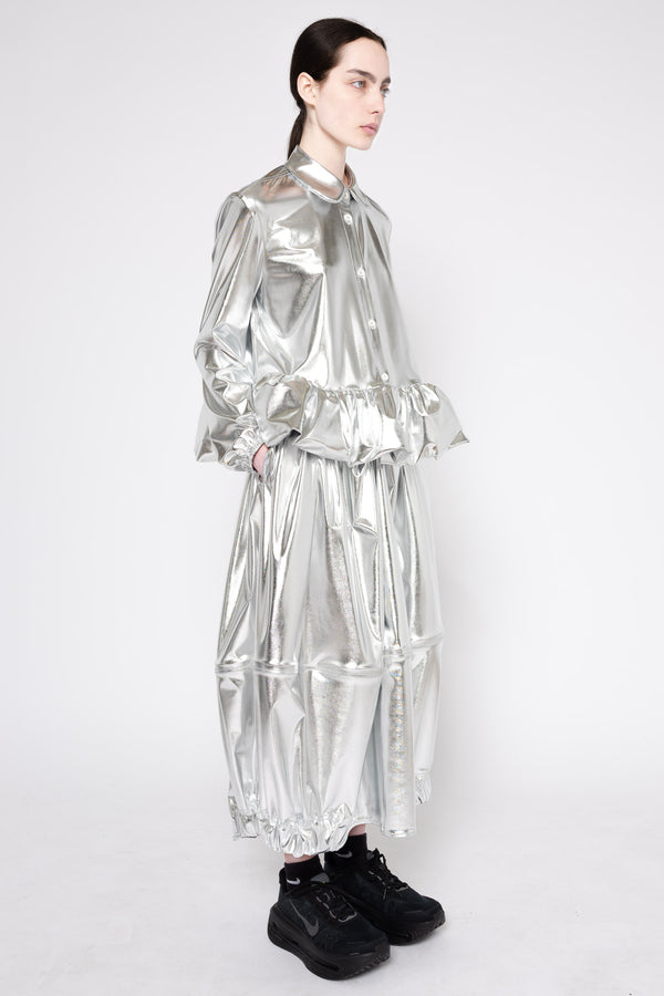A model wearing a shiny metallic silver long-sleeve dress with a high collar, gathered waist, and voluminous tiered skirt, paired with black sneakers, against a plain light gray background.