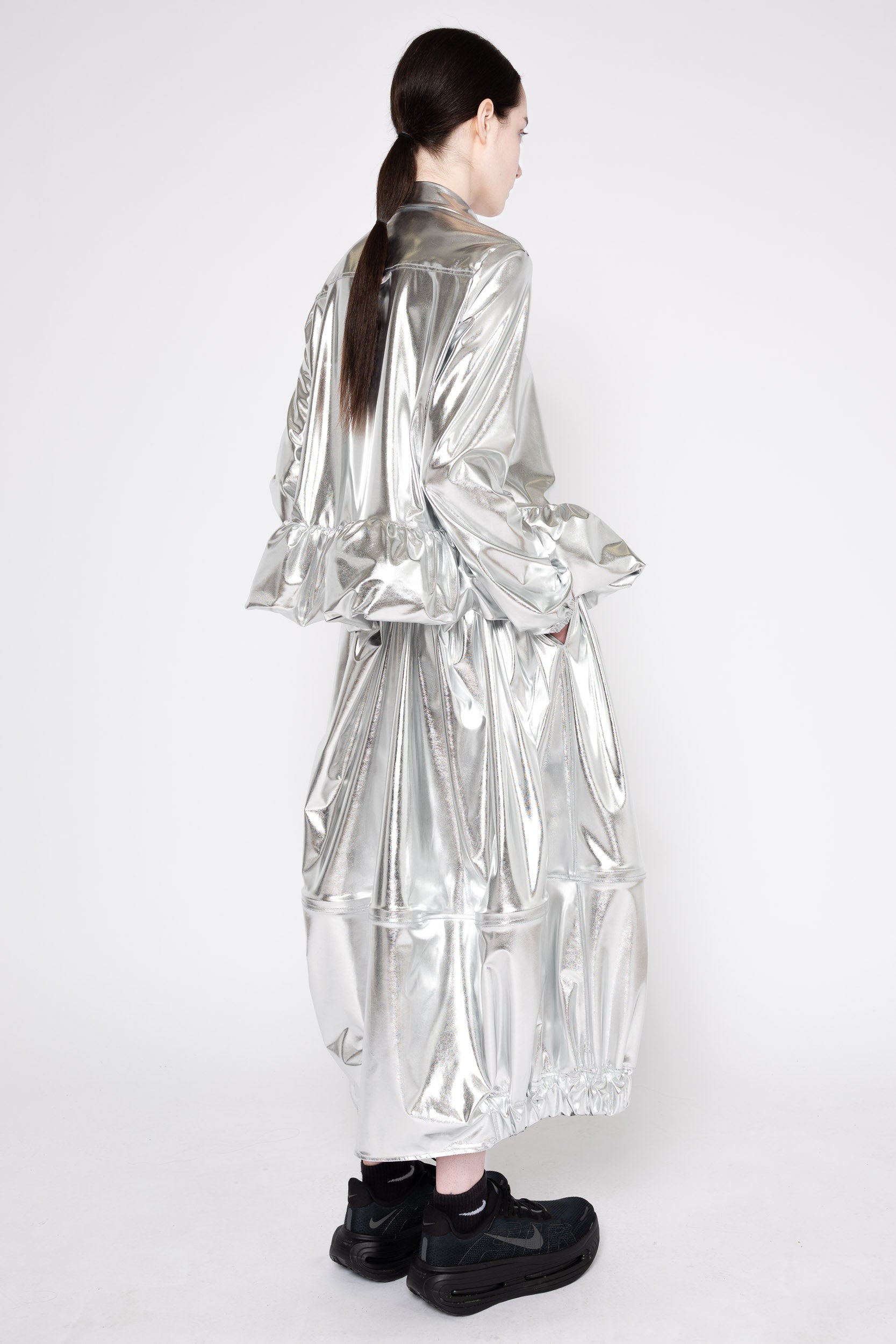A model shown from the back wearing a long, shiny silver metallic coat with voluminous ruffled sleeves and a tiered skirt, paired with black shoes, against a plain white studio background.