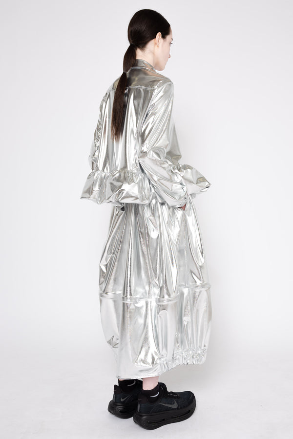 A model shown from the back wearing a long, shiny silver metallic coat with voluminous ruffled sleeves and a tiered skirt, paired with black shoes, against a plain white studio background.