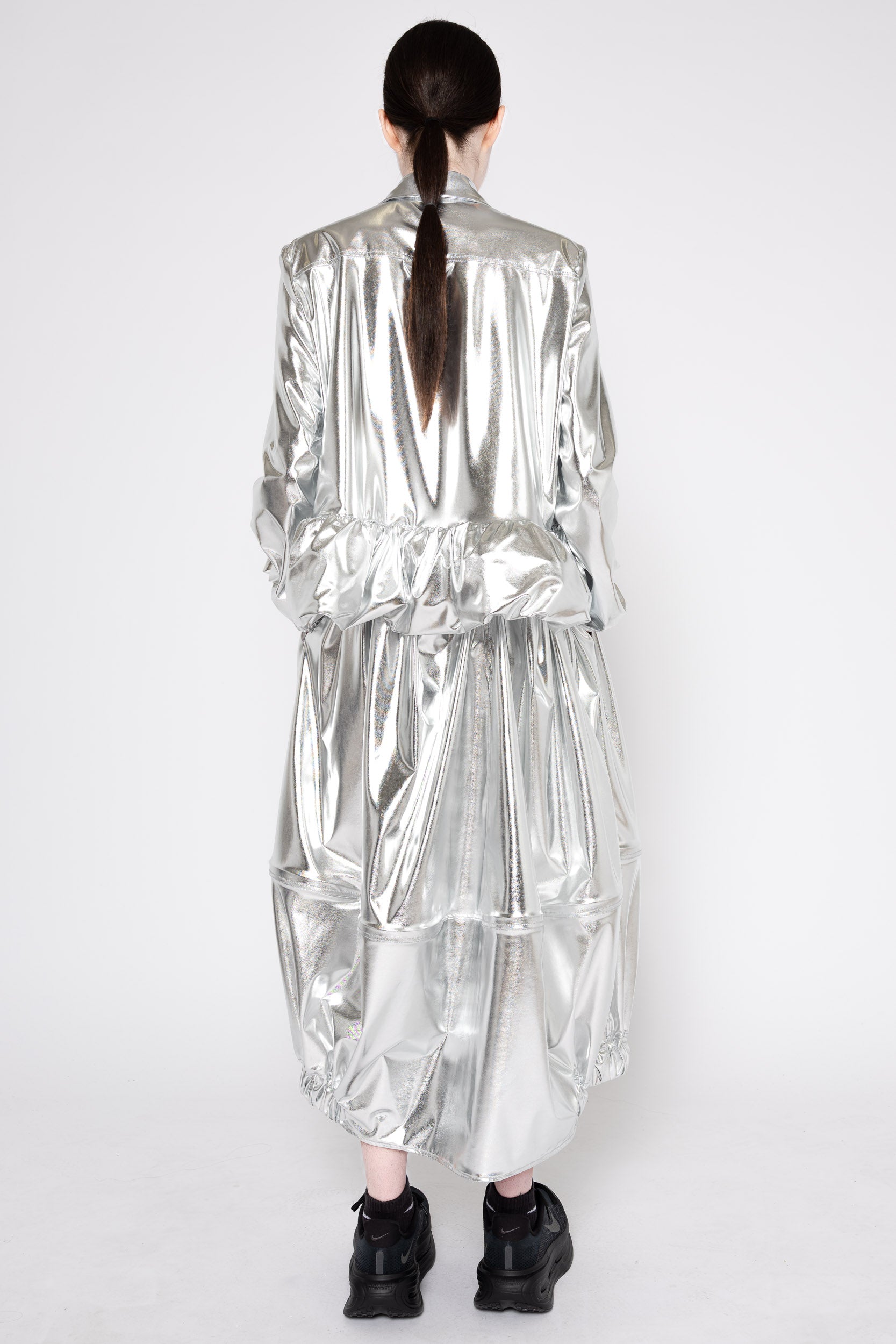 A model viewed from the back wearing a voluminous, metallic silver jacket and matching pleated skirt, paired with black sneakers, against a plain white studio background.