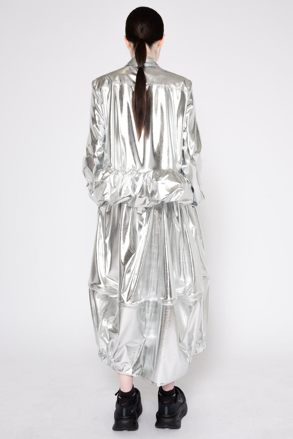 A model viewed from the back wearing a voluminous, metallic silver jacket and matching pleated skirt, paired with black sneakers, against a plain white studio background.