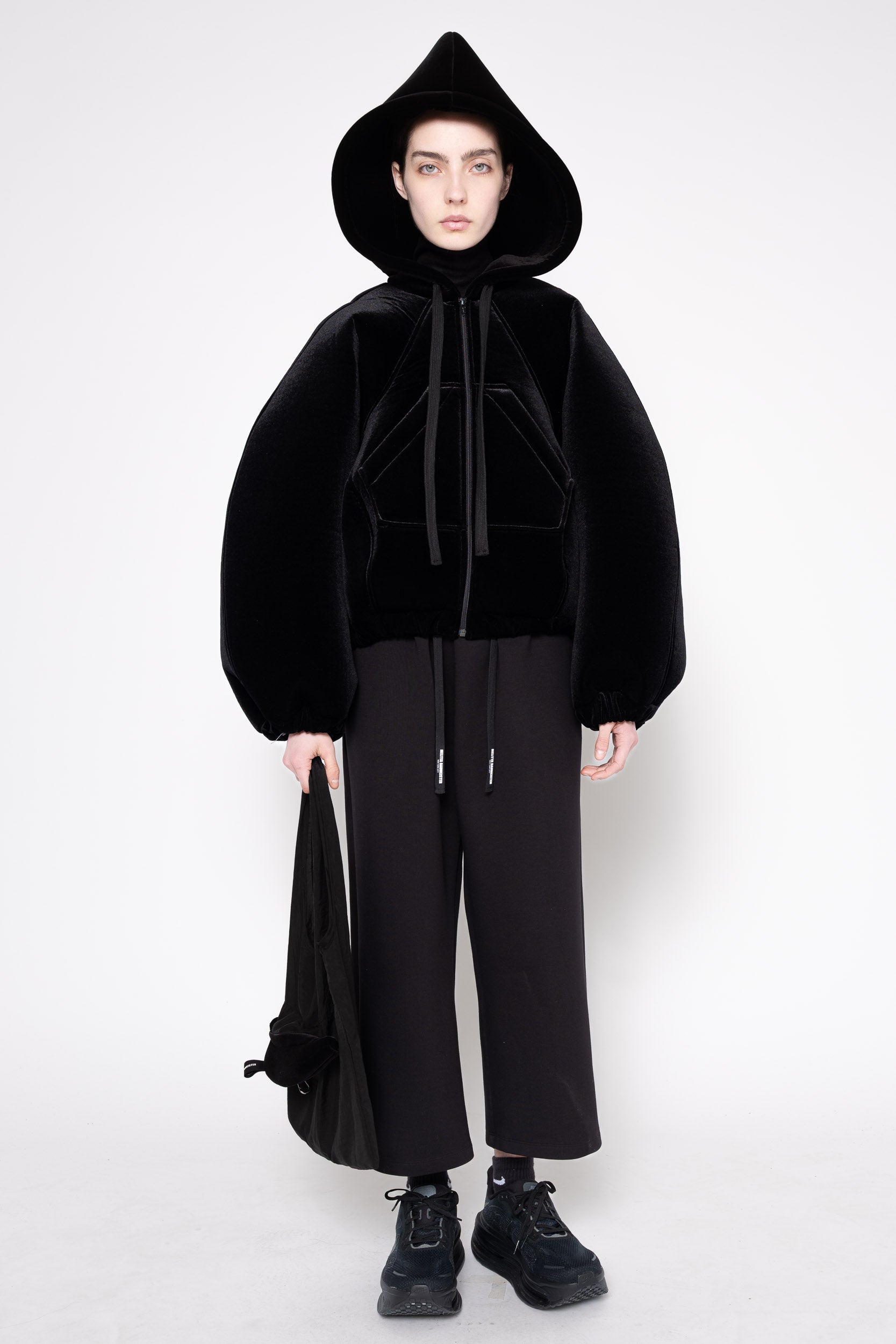 A model stands against a light gray studio background wearing a black hooded jacket with a fuzzy texture, black wide-leg pants, and black sneakers, while holding a black bag.